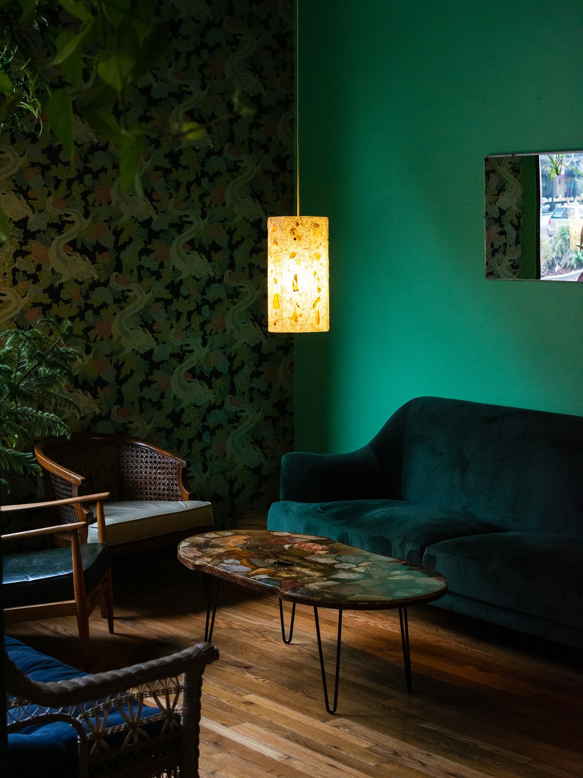 Living room corner with dark green botanical wallpaper, a teal velvet sofa and a glowing pendant lamp