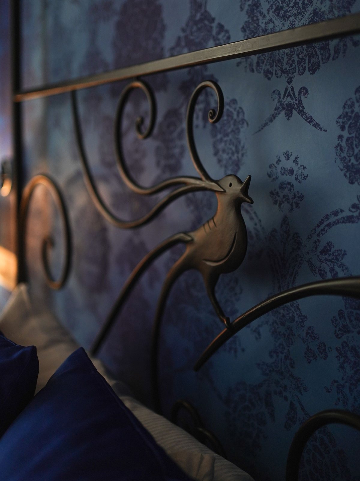Deep blue damask wallpaper behind a scrolled metal headboard with a bird motif
