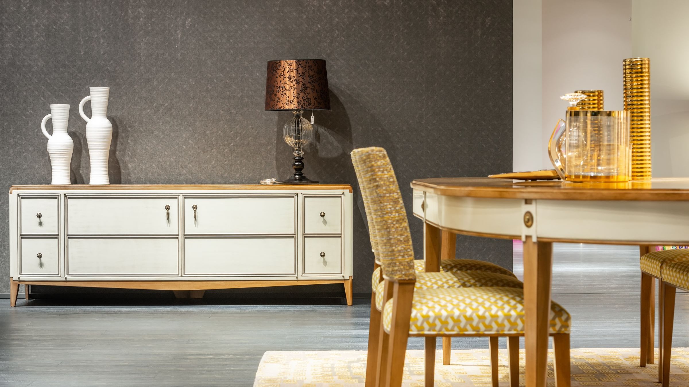 Dining room with deep charcoal patterned wallpaper behind a cream credenza, with a round wooden table and gold-toned glassware